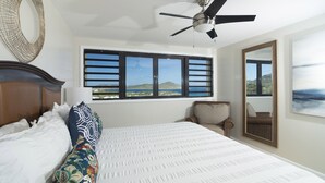 A bedroom with a large bed, a ceiling fan, a window with a view of hills, a chair, and a mirror.