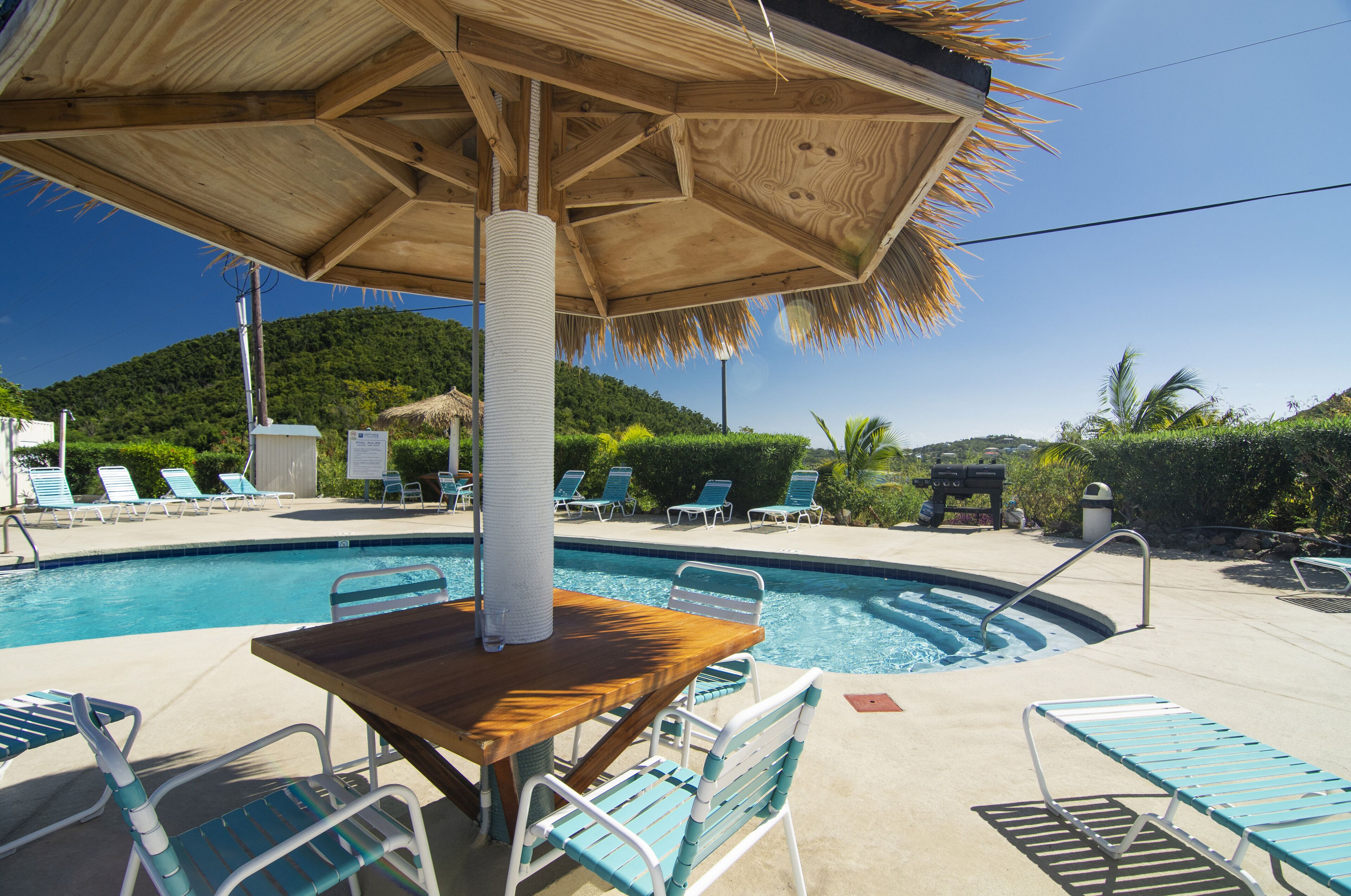 A pool area with lounge chairs, a wooden table, and a thatched umbrella.