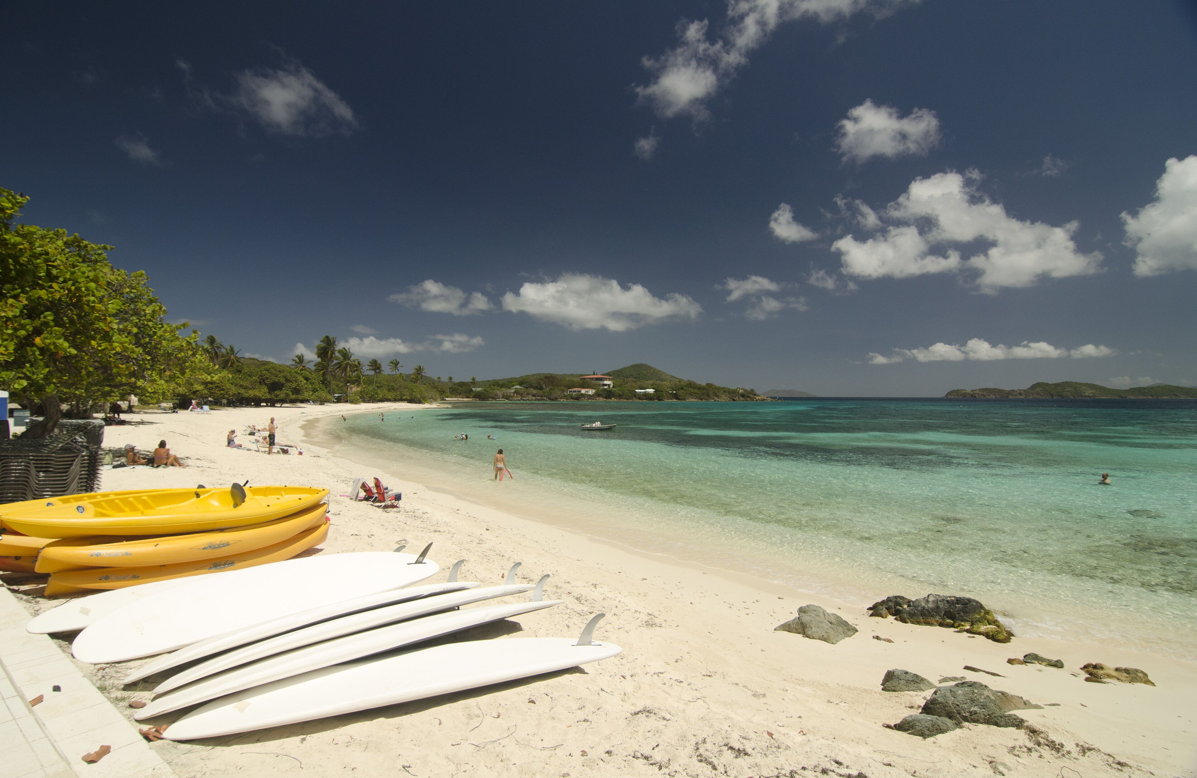 Een strand met surfplanken en kayaks, helder turquoise water, en enkele mensen in de verte.