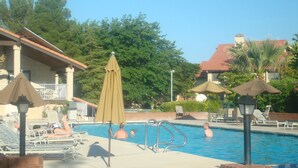 A swimming pool with lounge chairs and umbrellas, surrounded by trees and a building with a red roof. - Great Deck for Spectacular Sunrises in Sports Village (St George)