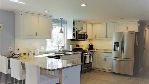 A modern kitchen with white cabinets, stainless steel appliances, a central island with bar stools, and a large window providing natural light. - Fabulous 3BR/3BA vacation house in Cotuit that sleeps 8 and only 1 mile to beach (Cotuit)