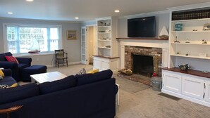 A living room with a fireplace, a television mounted above it, and a bookshelf with decorative items. - Fabulous 3BR/3BA vacation house in Cotuit that sleeps 8 and only 1 mile to beach (Cotuit)