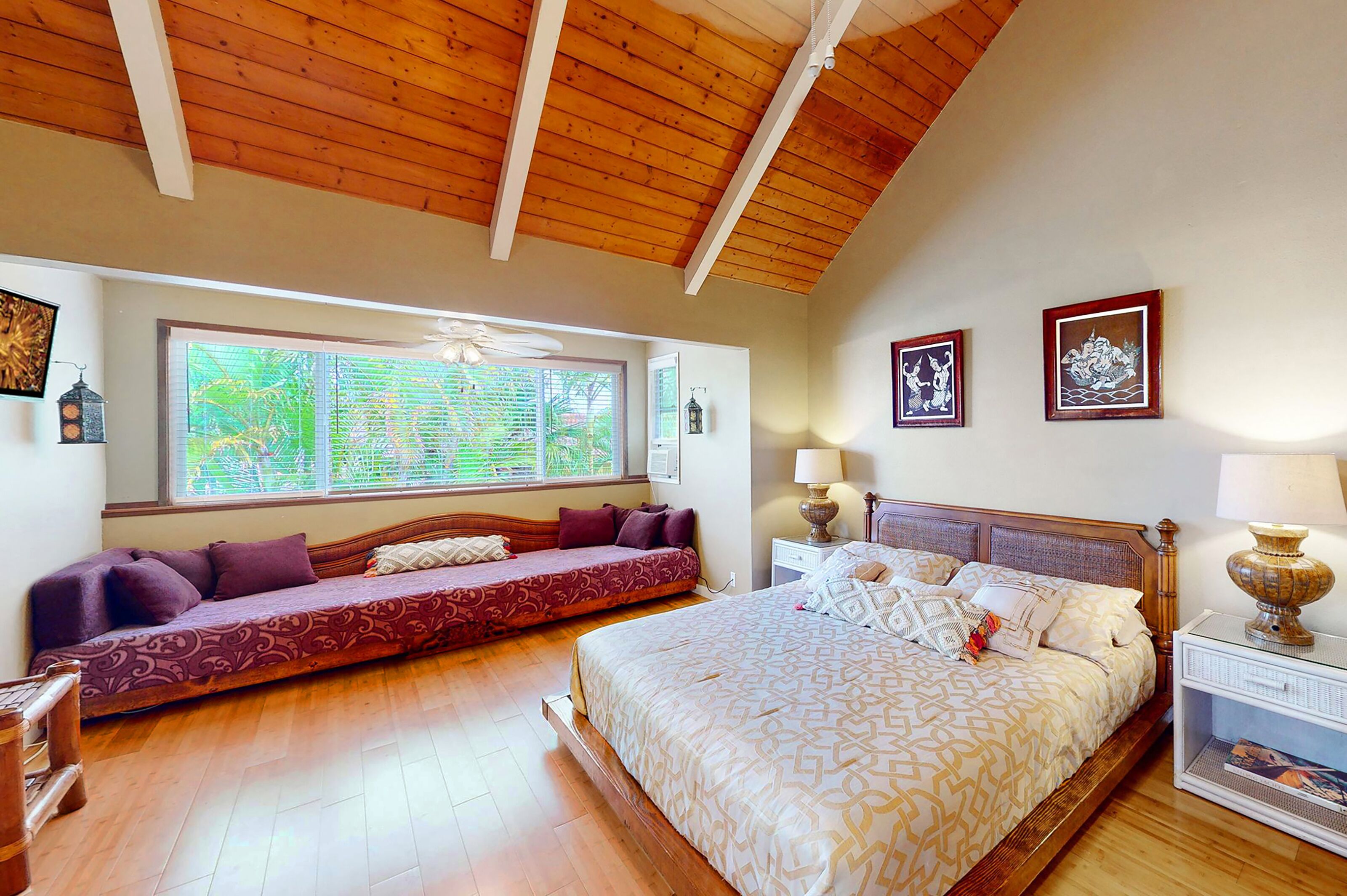 A bedroom with a large bed, a sofa bed, wooden flooring, a ceiling fan, and a window with blinds.
