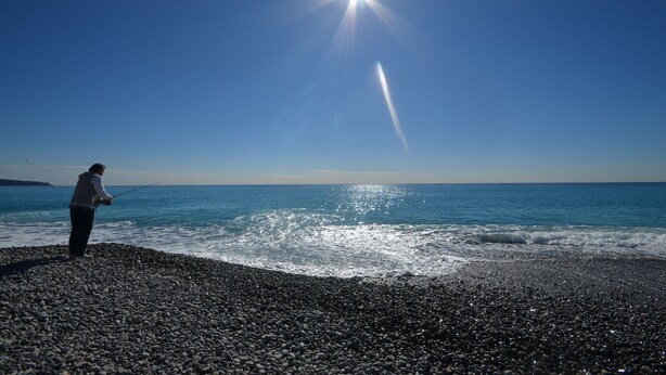 Una persona pesca a una platja de galets sota un cel serè i amb el sol resplendent.
