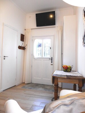 Een kamer met een witte deur, een televisie erboven, een klein tafeltje met een fruitmand en een houten stoel.