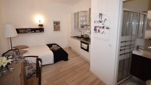 Una habitació petita i funcional amb llit, taula de nit i cadira. A prop hi ha una cuineta amb rentatlla, fogons i armaris d'emmagatzematge. Es veu un bany petit amb vàter i rentatlla.