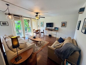 A living room with a sofa, coffee table, and TV stand. There is a balcony with a view of greenery.