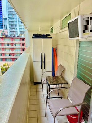 A balcony with a view of a building, a white wardrobe, two chairs, and an air conditioning unit.