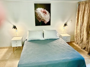 A bedroom with a bed, two bedside tables, a nightstand, a wall-mounted lamp, and a window with curtains.