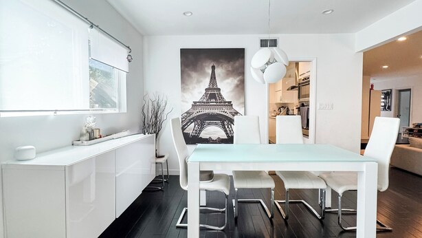 A modern kitchen with a dining area, featuring a large window, white cabinetry, a glass table, and a black and white Eiffel Tower painting on the wall. - Modern 5bdr House with hot tub 7 min from Universal studios (north hollywood)