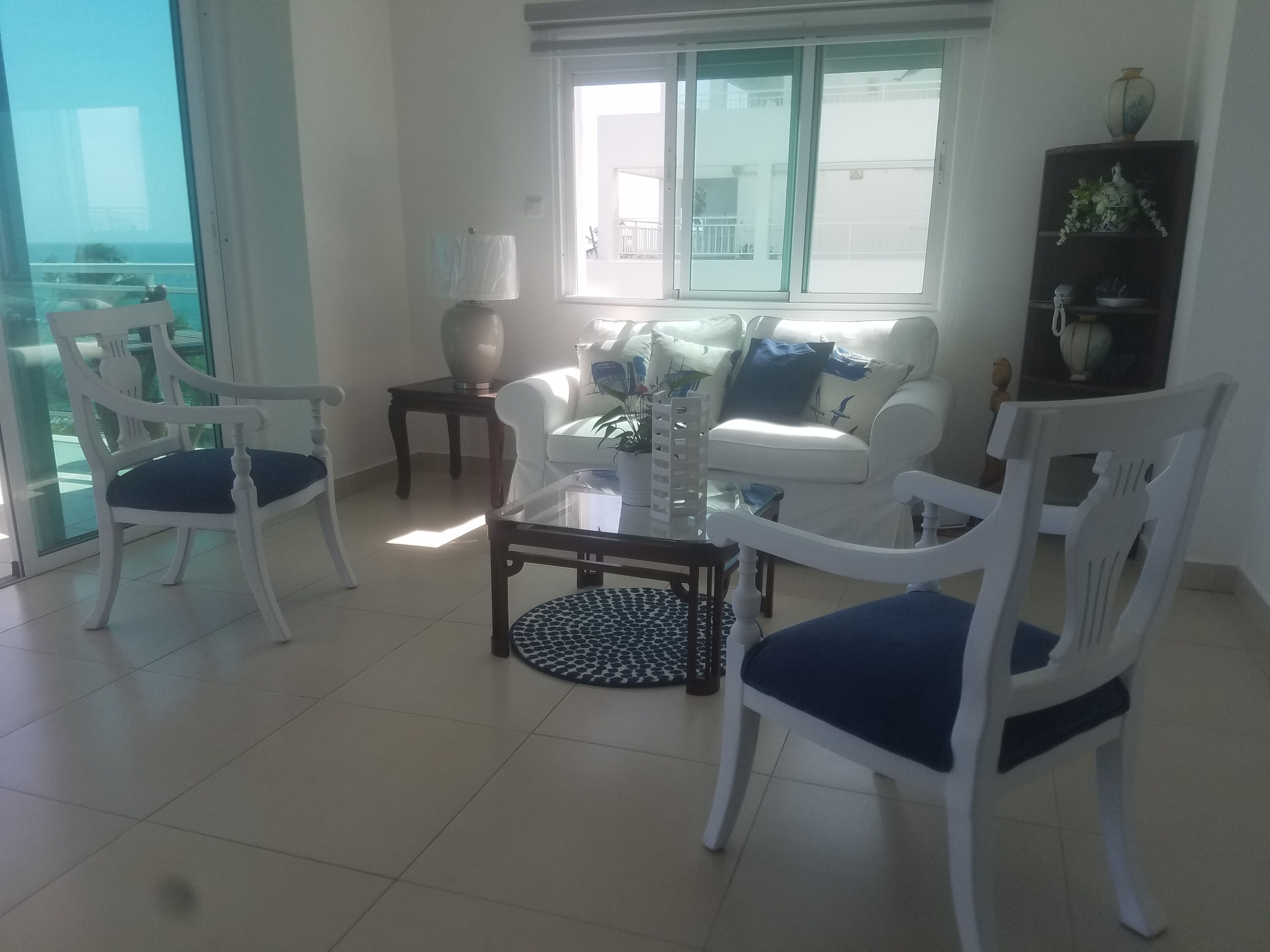 Un salon avec un canapé blanc, deux fauteuils blancs, une table basse en verre et une petite table d’appoint avec une lampe.