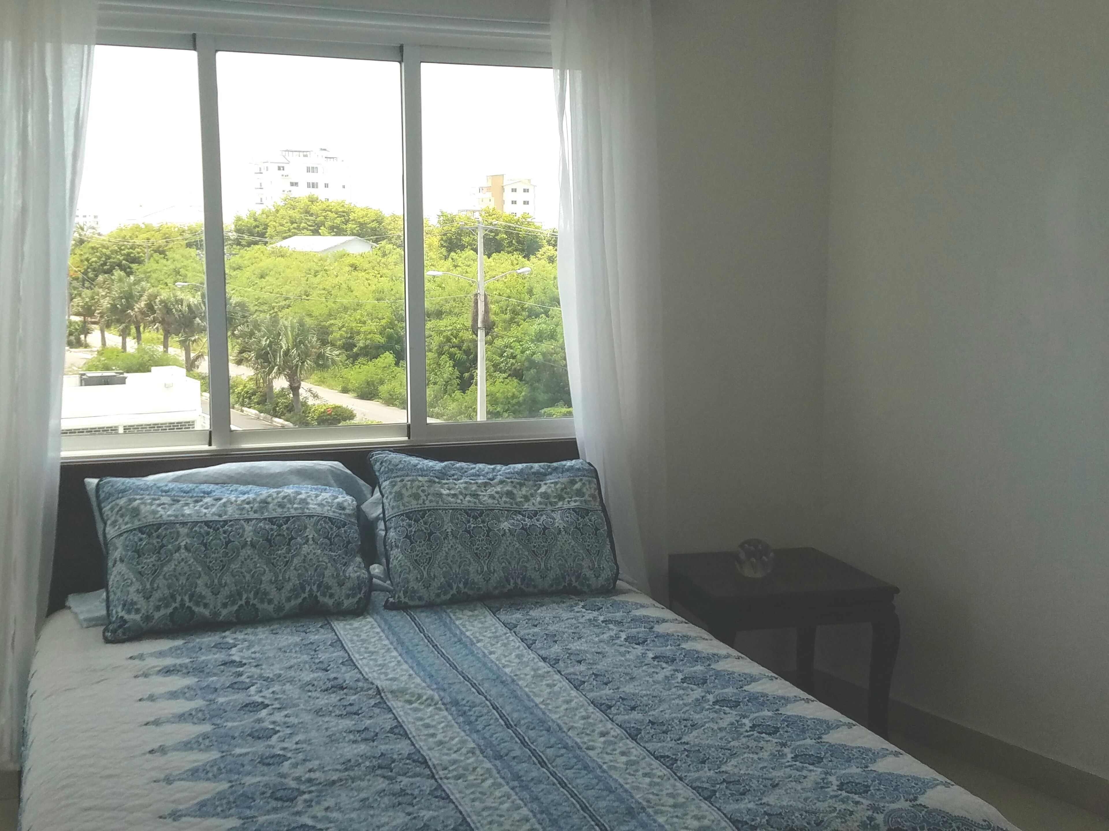 A bedroom with a large window, a bed with patterned bedding, and a small bedside table.