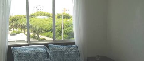 A bedroom with a large window, a bed with patterned bedding, and a small bedside table.