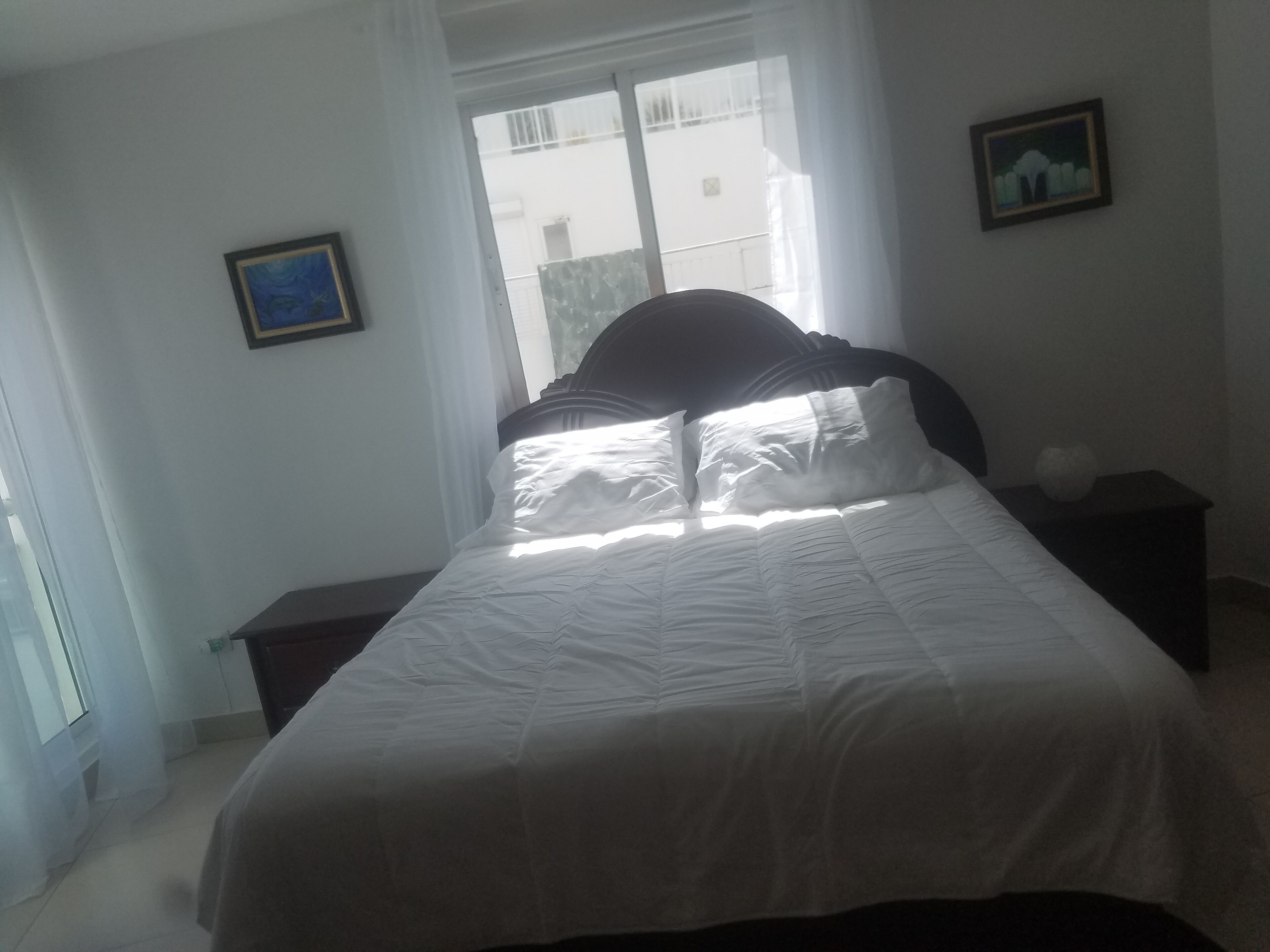 Een slaapkamer met een bed, nachtkastje, raam en twee ingekaderde foto’s aan de muur.