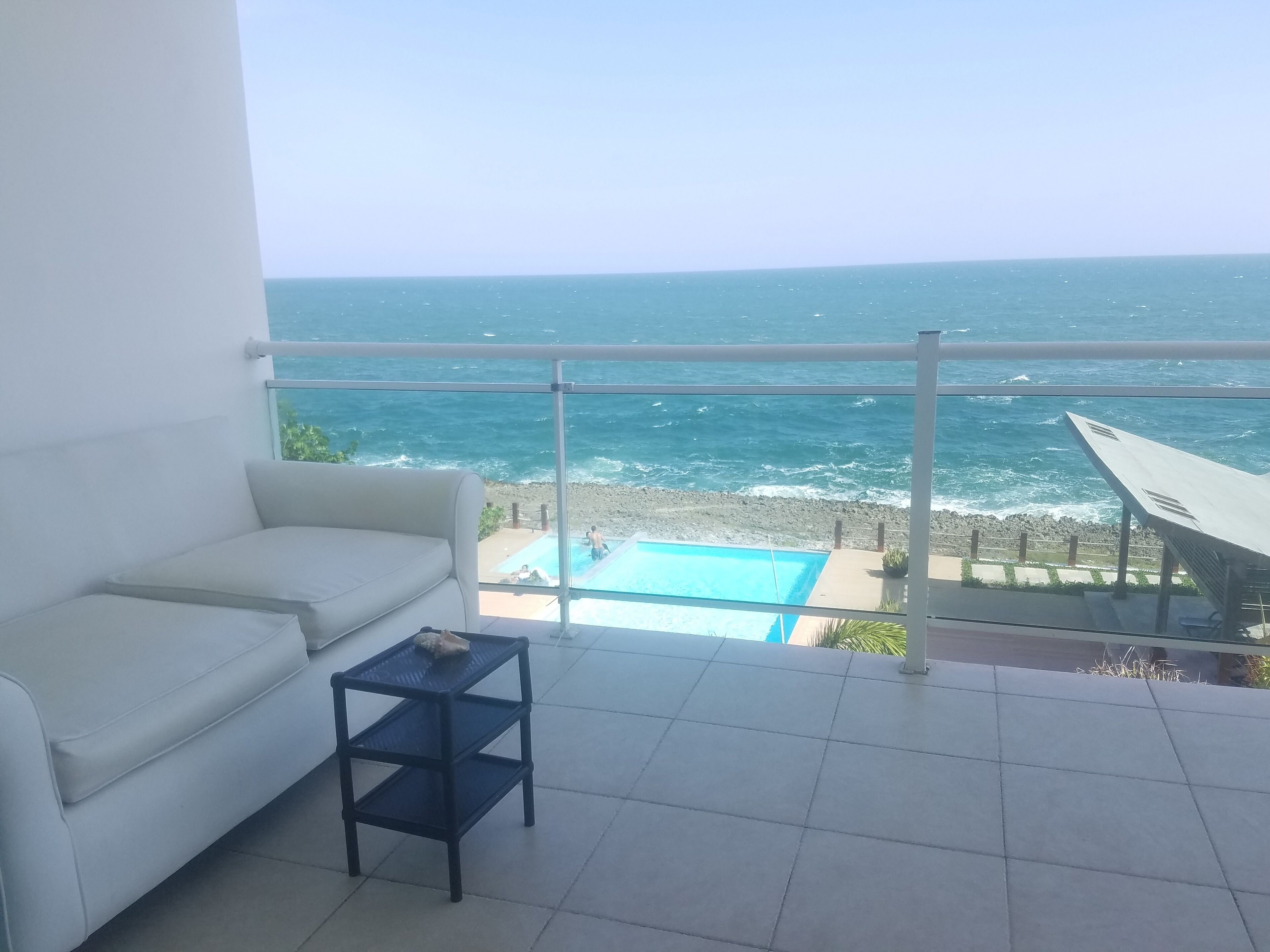 A balcony with a white sofa, a small table, and a view of the ocean and beach.