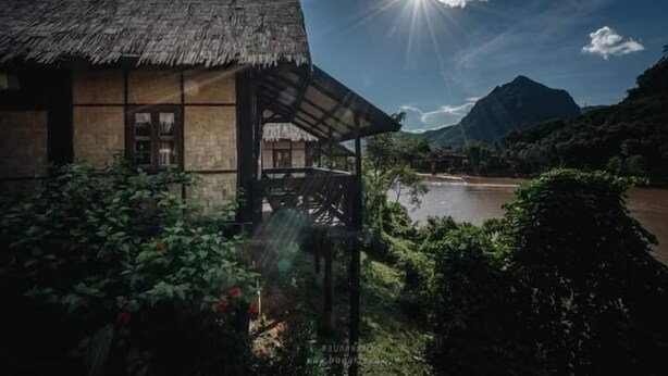 A rustic house with a thatched roof, a wooden porch, and a view of a river and mountains in the background. - Nong Kiau Riverside (Nong Khiaw)