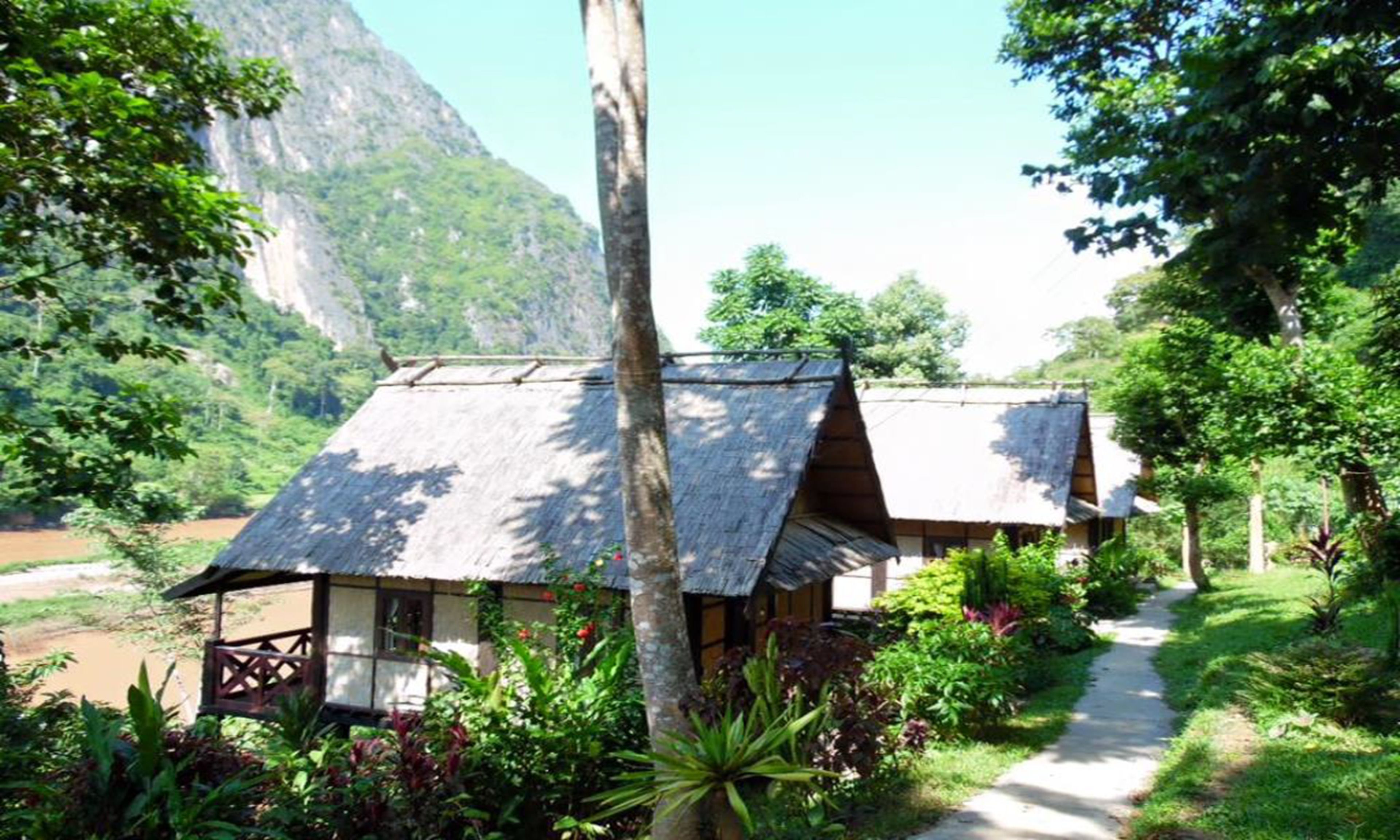 A traditional wooden house with a thatched roof, surrounded by lush greenery and a mountain in the background.