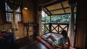 A room with a wooden floor, a bed, a window with curtains, and a view of greenery outside. - Nong Kiau Riverside (Nong Khiaw)