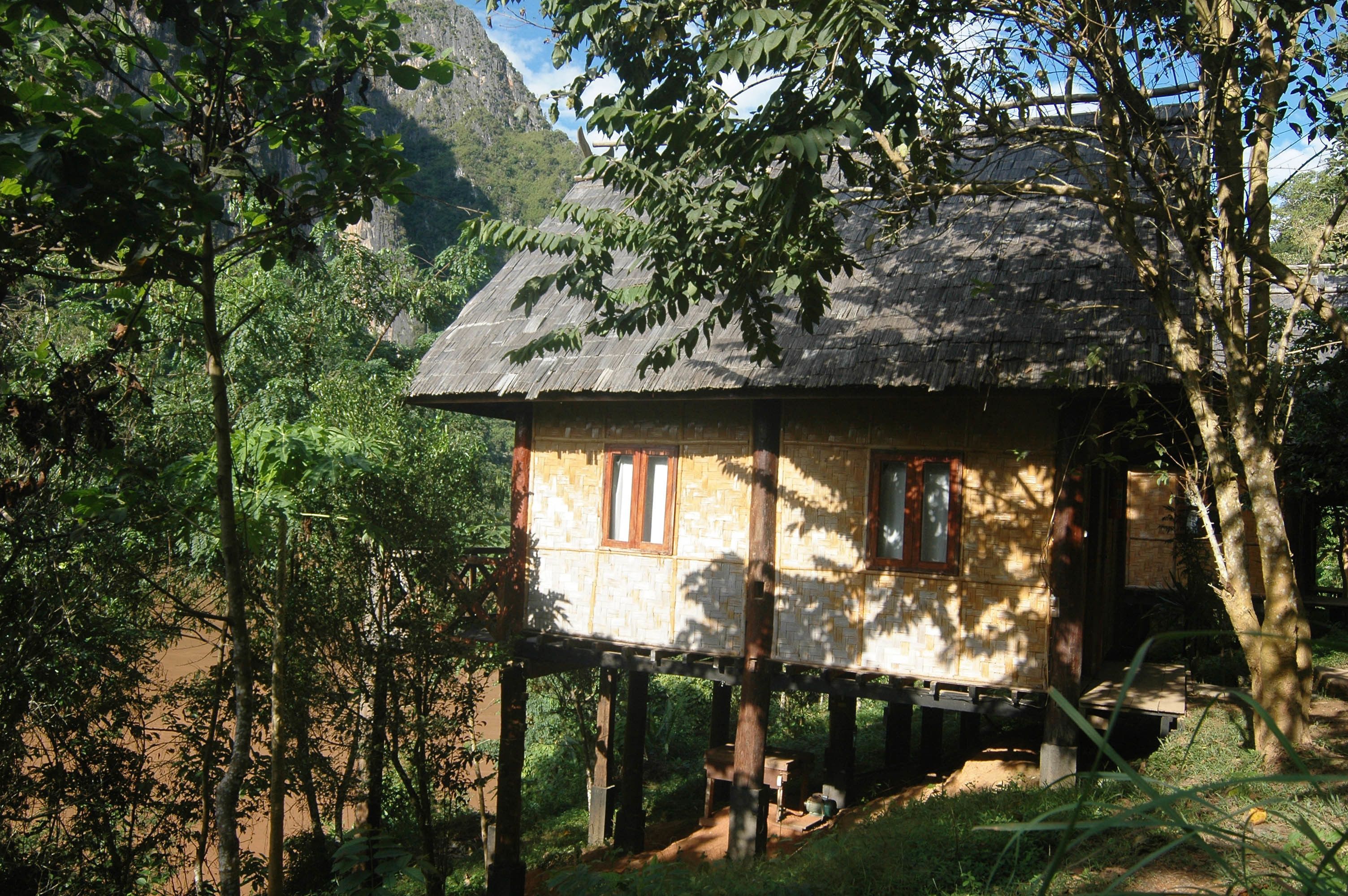 A rustic wooden house elevated on stilts, surrounded by dense forest and mountains.