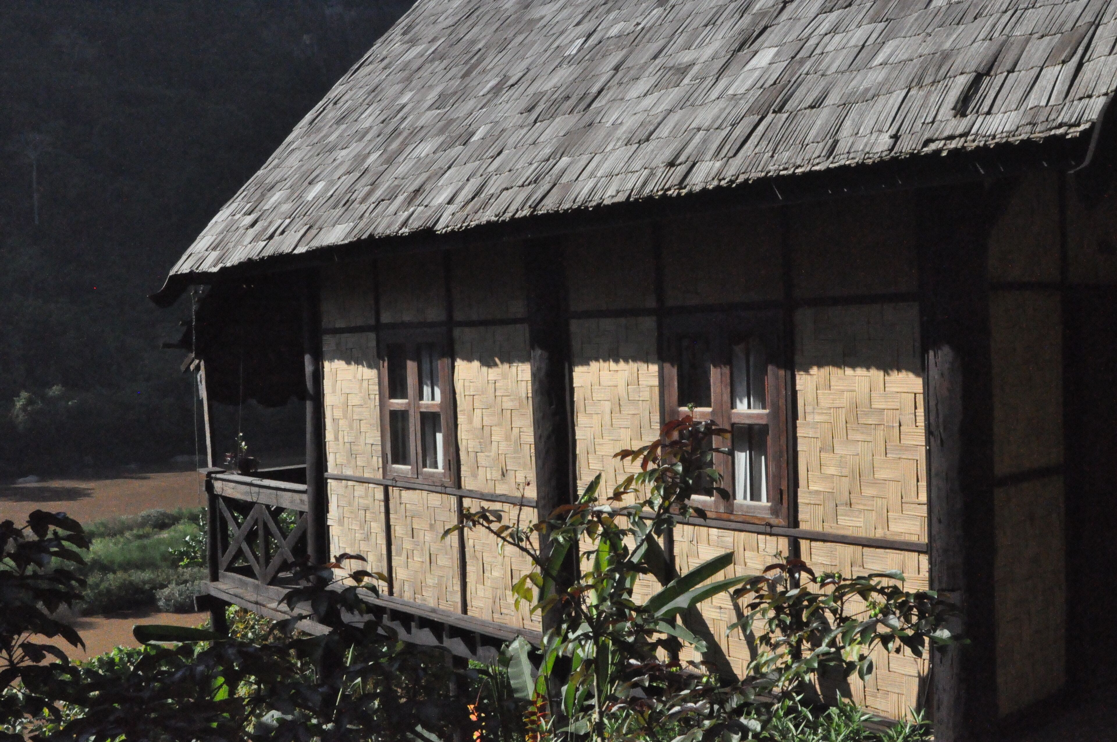 A rustic wooden house with a thatched roof, a wooden balcony, and a woven wall.