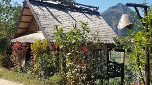 A rustic hut with a sloped roof, surrounded by lush greenery and a mountain backdrop. - Nong Kiau Riverside (Nong Khiaw)