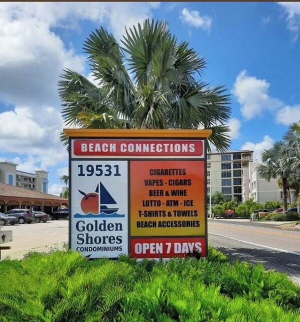 Letrero de 'Golden Shores Condominiums' publicitando 'Beach Connections' con servicios como cigarrillos, vapeadores, cerveza, vino, lotería, cajero automático, hielo, camisetas, toallas y artículos para la playa, abierto 7 días a la semana.