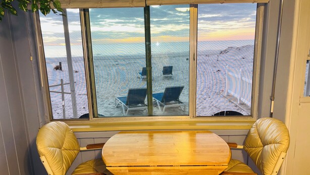 A room with a large window overlooking a beach, a round wooden table, and two leather chairs.