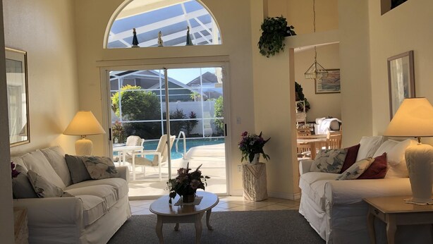 Un salón amplio con dos sofás blancos, una mesa de centro y vistas a una piscina a través de grandes ventanales arqueados.