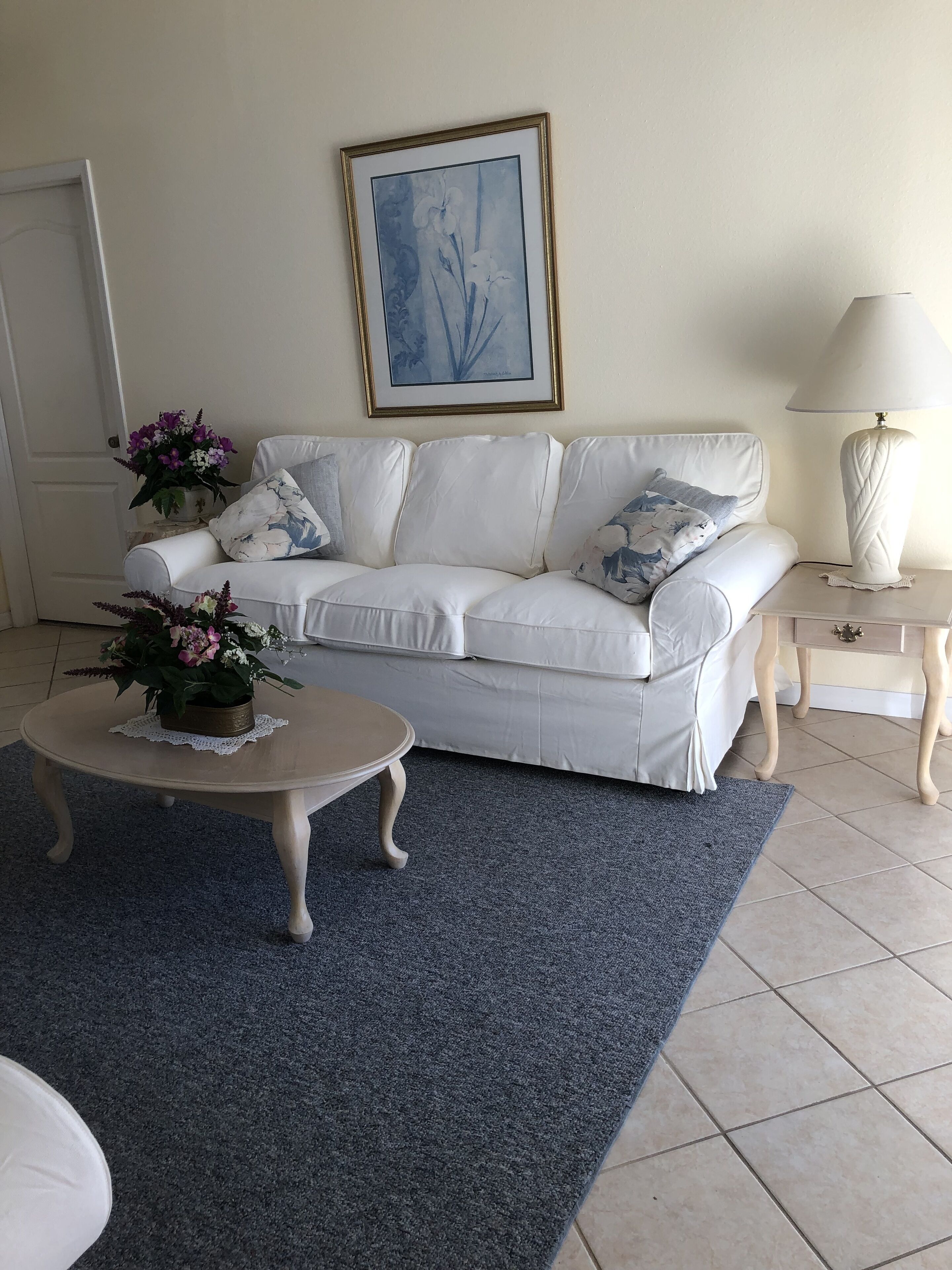 Een woonkamer met een witte bank, een ronde salontafel met een bloemdecoratie, een kunstwerk in een lijst aan de muur, en een witte lamp op een bijzettafeltje.