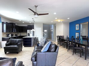 A modern living room with a leather sofa, a dining area with a table and chairs, a kitchen with dark cabinets and appliances, and a ceiling fan.