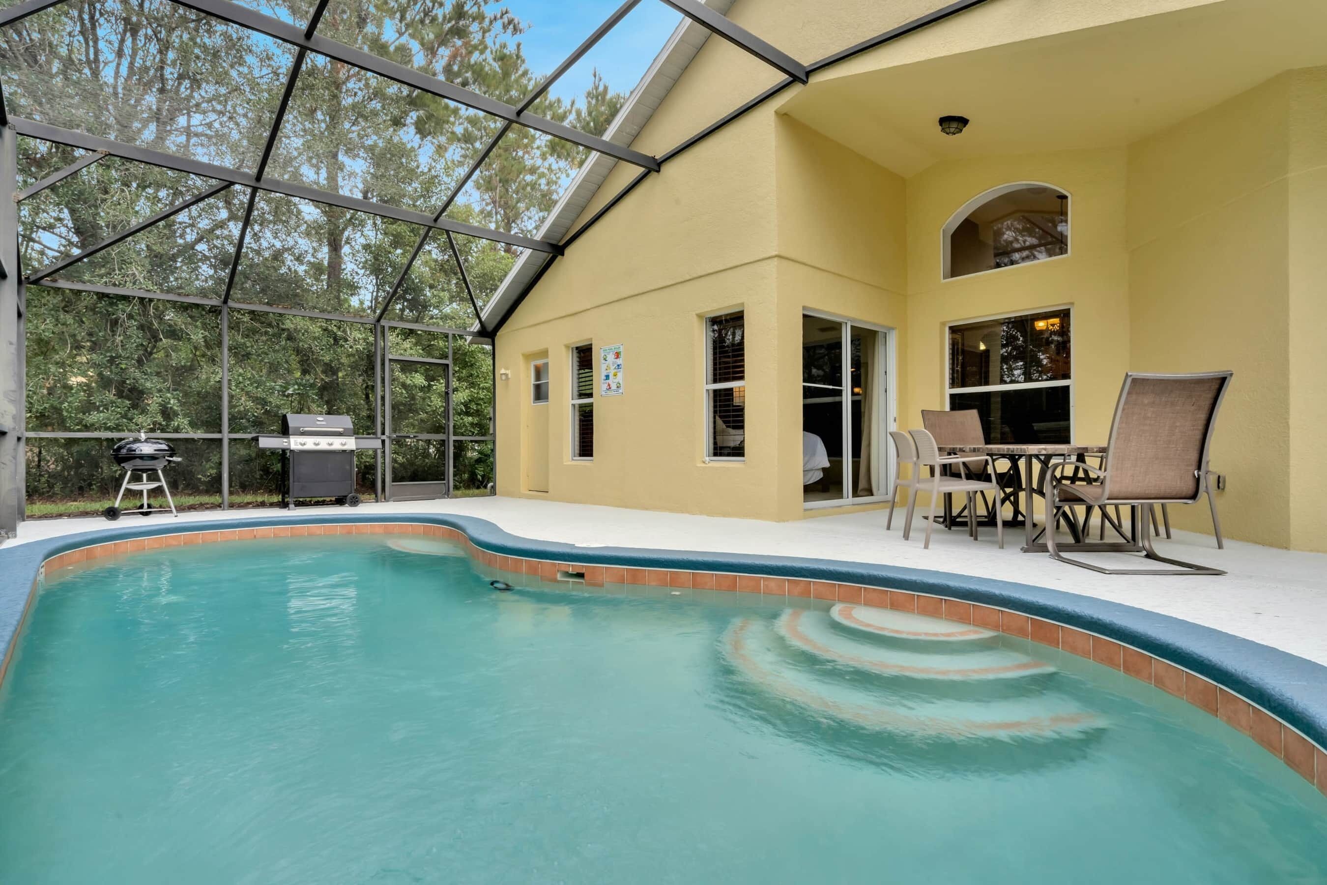 Una piscina con recinto in vetro, un barbecue e area salotto all'aperto.