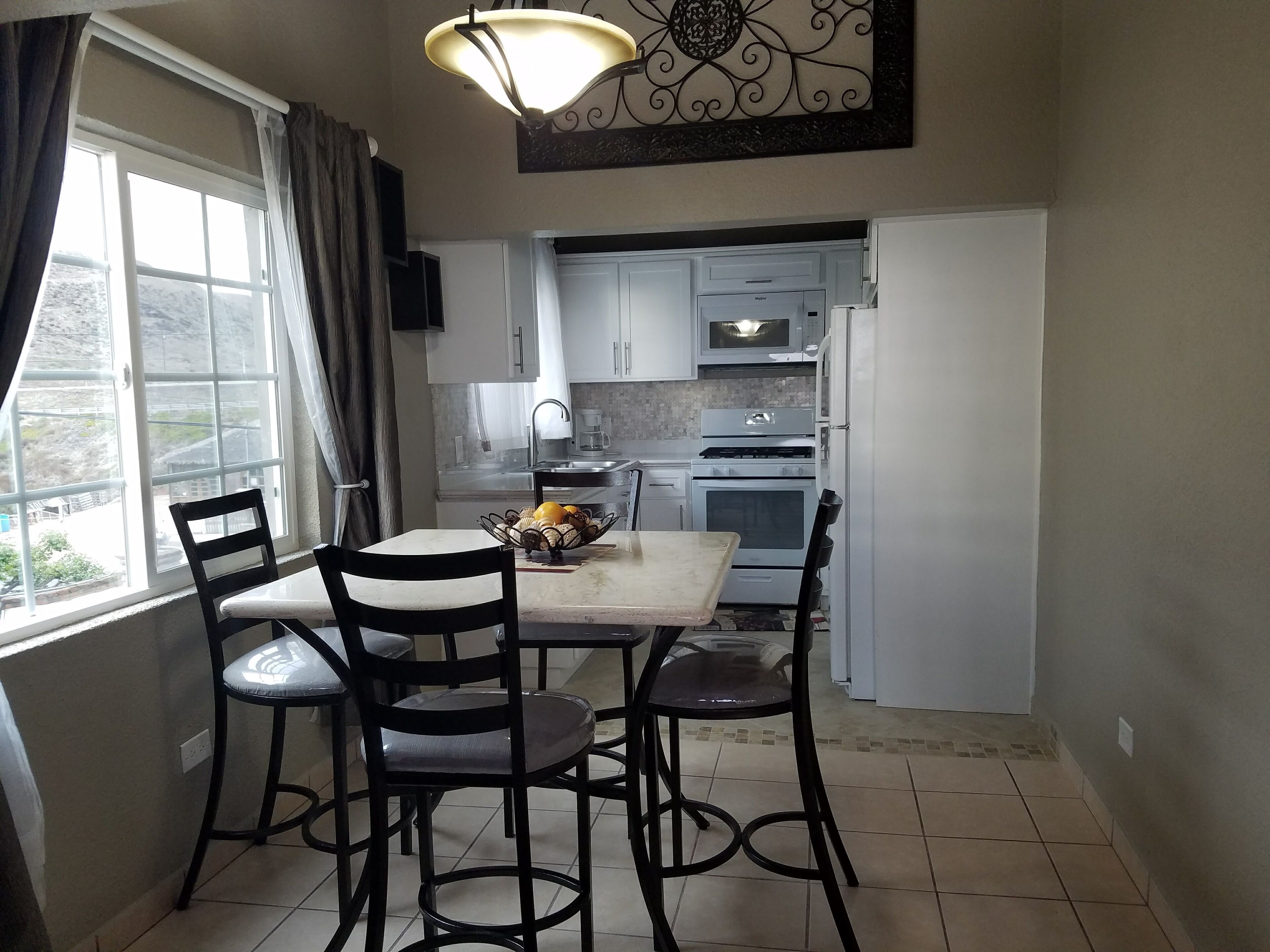 A compact kitchen with white cabinets, a microwave, an oven, and a sink. There is a dining table with four chairs and a fruit bowl on it.