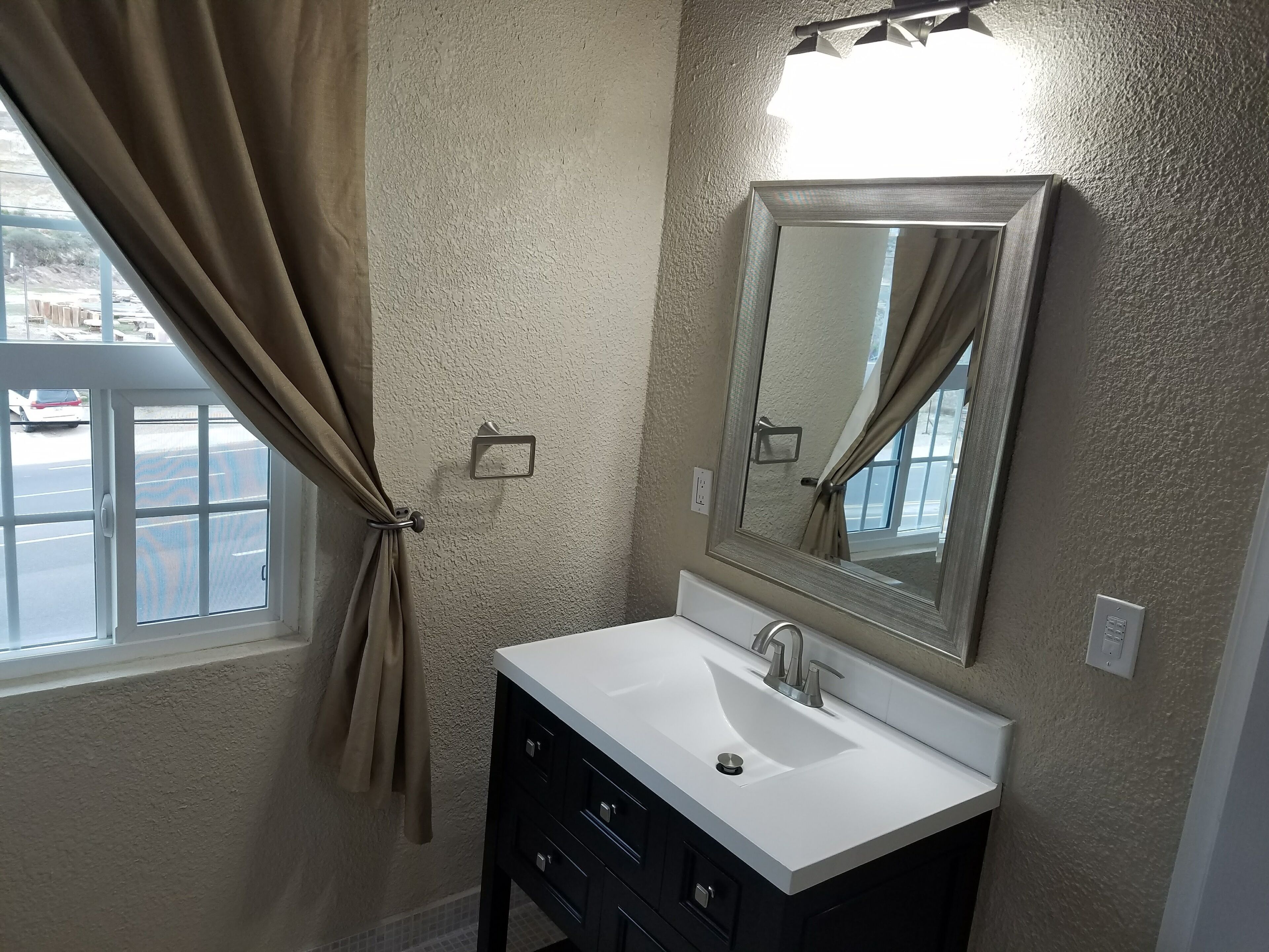 A bathroom with a white sink, dark cabinets, a large mirror, and a window with curtains.