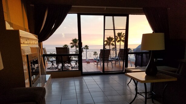 Une chambre avec vue sur le coucher de soleil sur l’océan, un balcon avec des chaises et une table avec une lampe.