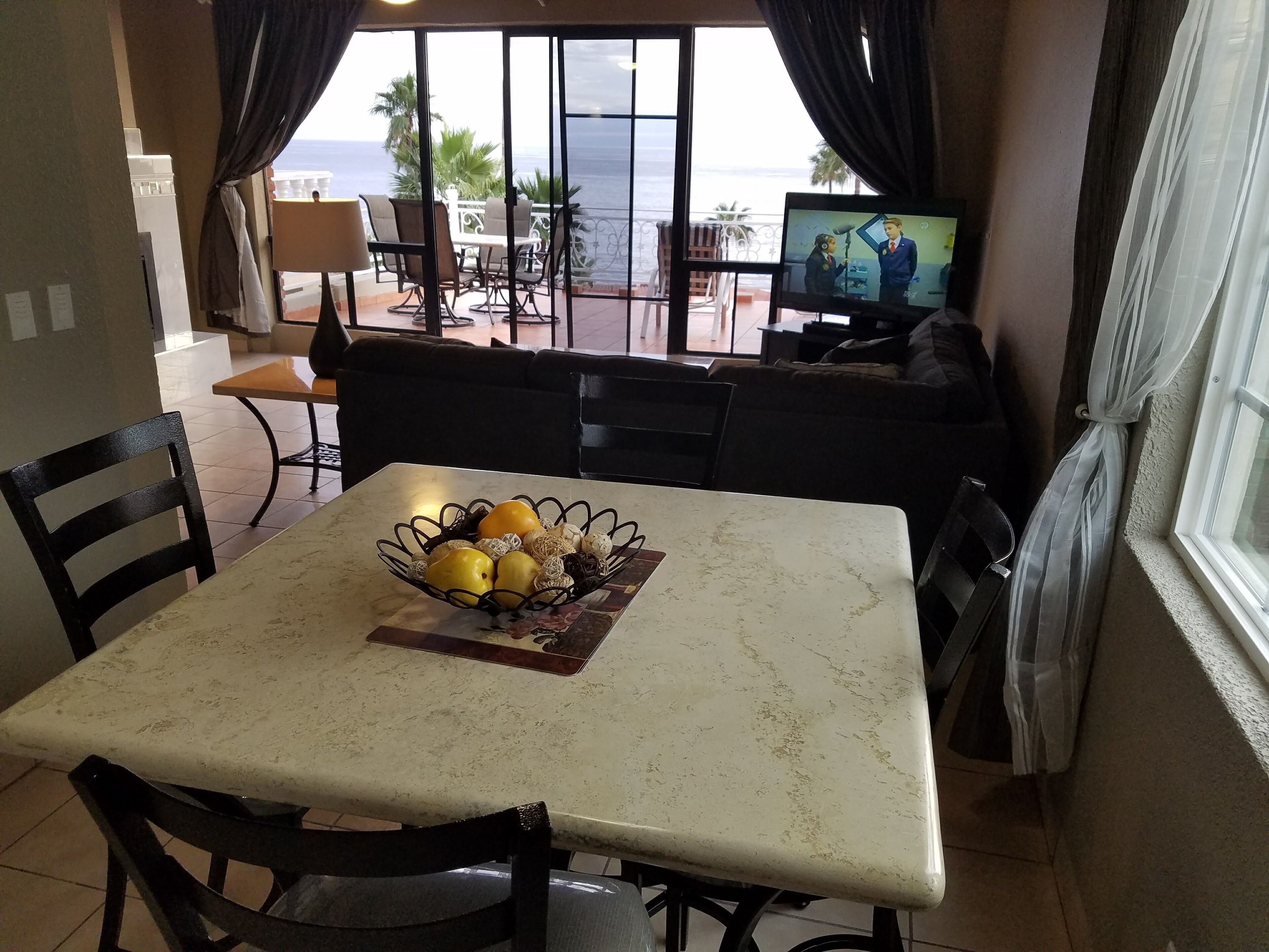Un comedor con una mesa lista para dos personas, un cuenco de fruta y un televisor al fondo.