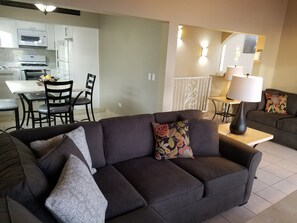 A modern living room with a dark sofa, a dining area with a white table and chairs, and a kitchen with white appliances.
