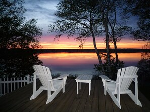 A wooden deck with two Adirondack chairs and a small table, overlooking a calm lake at sunset.
