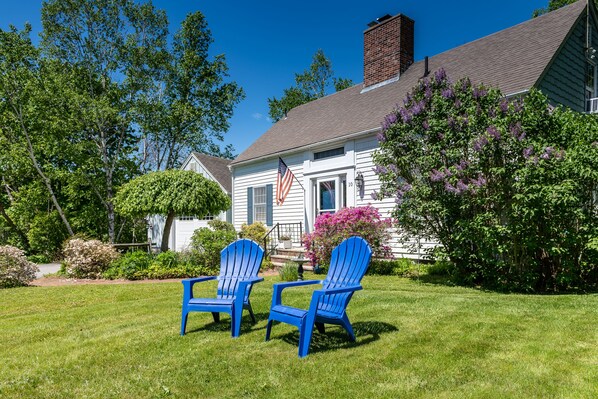 A house with a flag, two blue Adirondack chairs, and a well-maintained lawn.