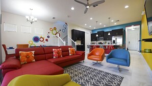 A modern living room with a sectional sofa, a yellow armchair, and a blue armchair. There is a kitchen in the background with dark cabinets and a stainless steel refrigerator. - EPIC VALUE-Disney Retreat! Magical Kid Friendly! Resort! (Davenport)