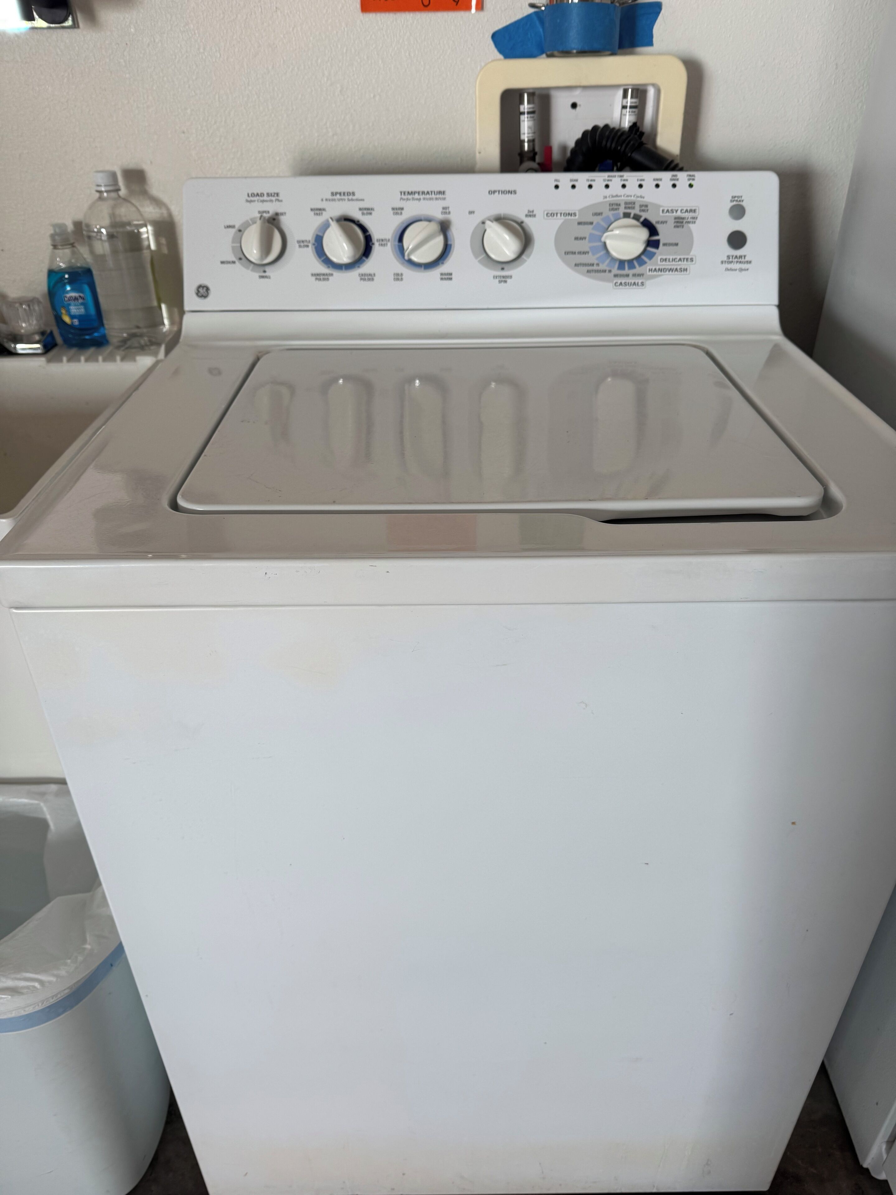 A front-loading washing machine with control panel, water bottles, and a blue towel on a white wall.