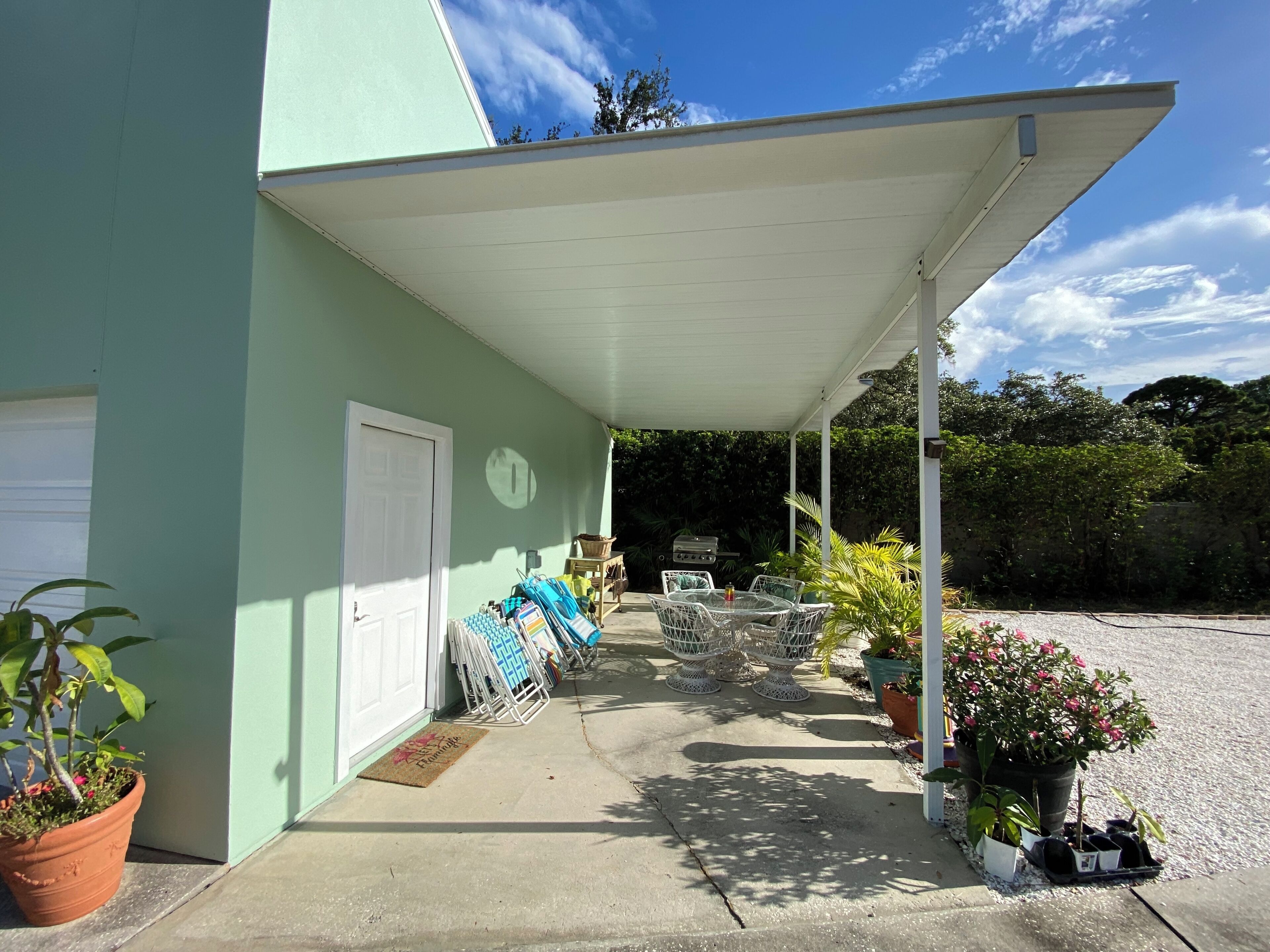 A covered outdoor area with a concrete floor, a white door, and a grill. There are several potted plants and umbrellas.