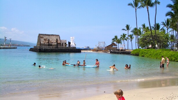A beach scene with people swimming and playing in the water, a child playing with sand on the shore, and palm trees in the background.