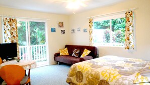 A bedroom with a bed, a sofa, a TV, and a chair. There are windows with curtains, a ceiling fan, and a view of greenery outside.