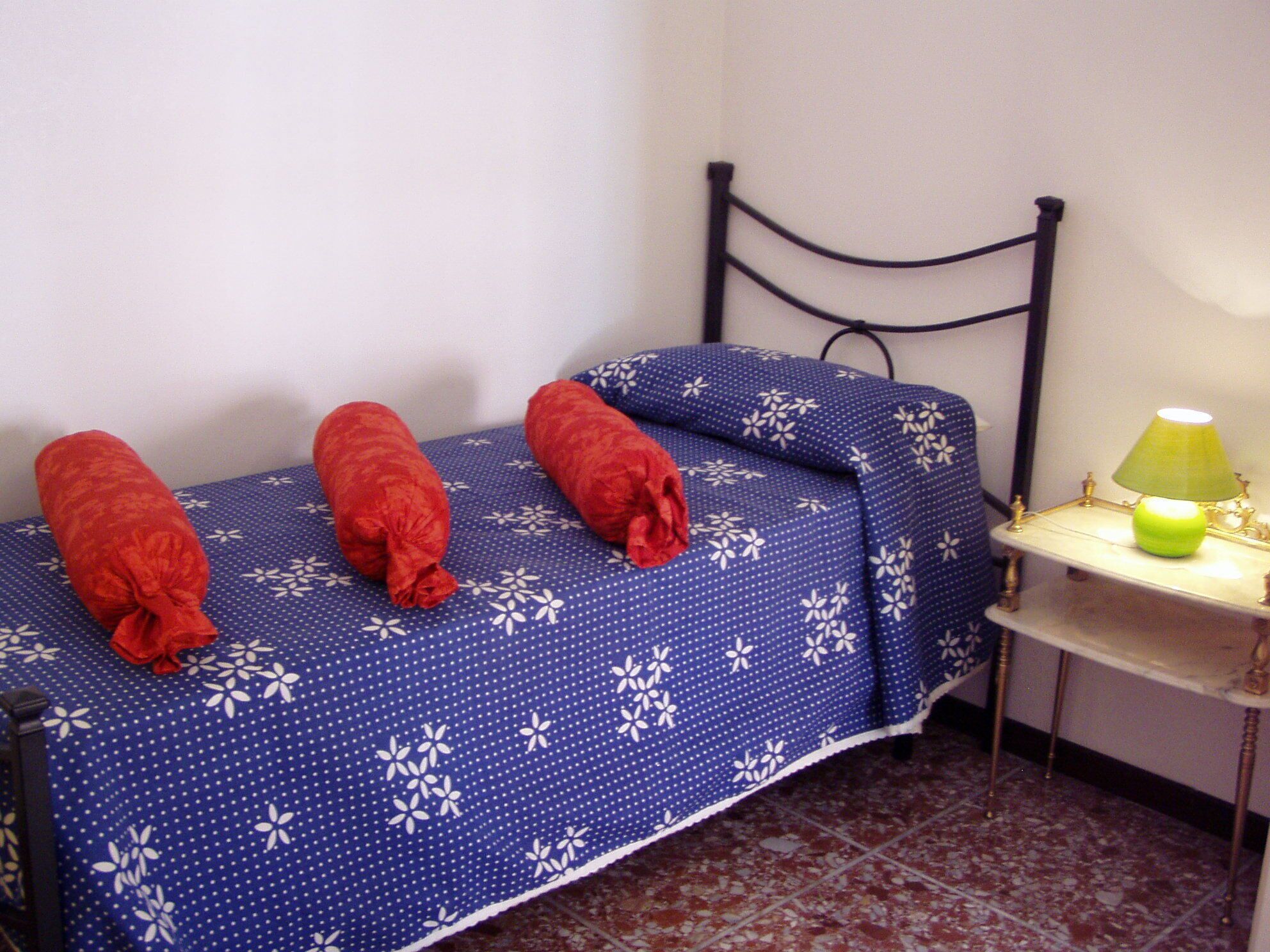 Una habitación pequeña con una cama cubierta por una colcha azul con motivos florales, una lámpara verde sobre una mesita de noche y una toalla roja en la cama.