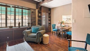 Una habitación acogedora con suelo de madera, un sillón verde, una silla azul, una mesita redonda y una estantería.