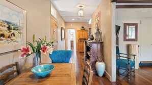 Un pasillo con una mesa de comedor de madera, sillas, un florero con flores, un sillón azul y un aparador de madera con objetos decorativos.