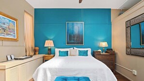 Un dormitorio con una cama grande, dos mesitas de noche, un taburete azul, un cuadro en la pared y un espejo en la pared opuesta.