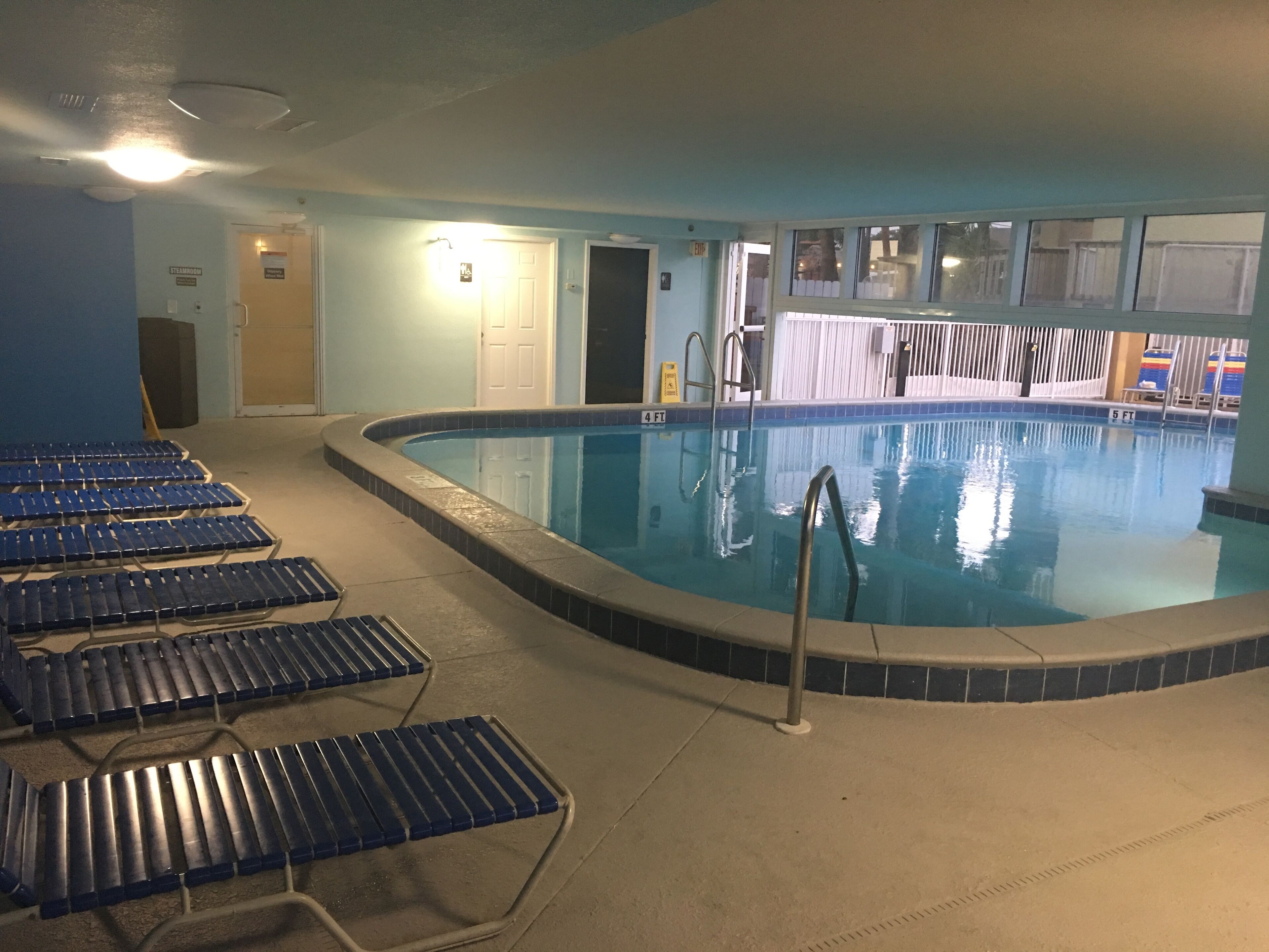 An indoor swimming pool with blue tiled walls, a pool ladder, and several empty lounge chairs.