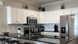 Una cocina moderna con armarios blancos, un refrigerador de acero inoxidable, un microondas y una cafetera. Hay tres taburetes negros y un bote de basura.