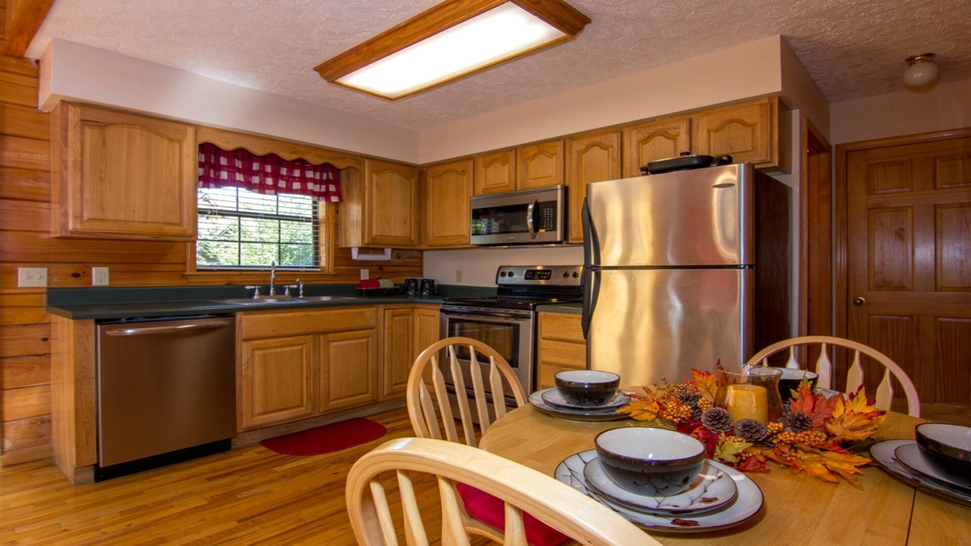 Une cuisine avec des meubles en bois, des appareils en acier inoxydable, une table à manger avec des chaises et une fenêtre avec des rideaux rouges.